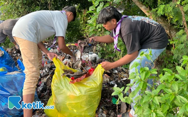 Foto Disaat Tengah sibuk memilah sampah di antara semak-semak, memasukkannya ke dalam kantong plastik besar berwarna kuning dan biru.